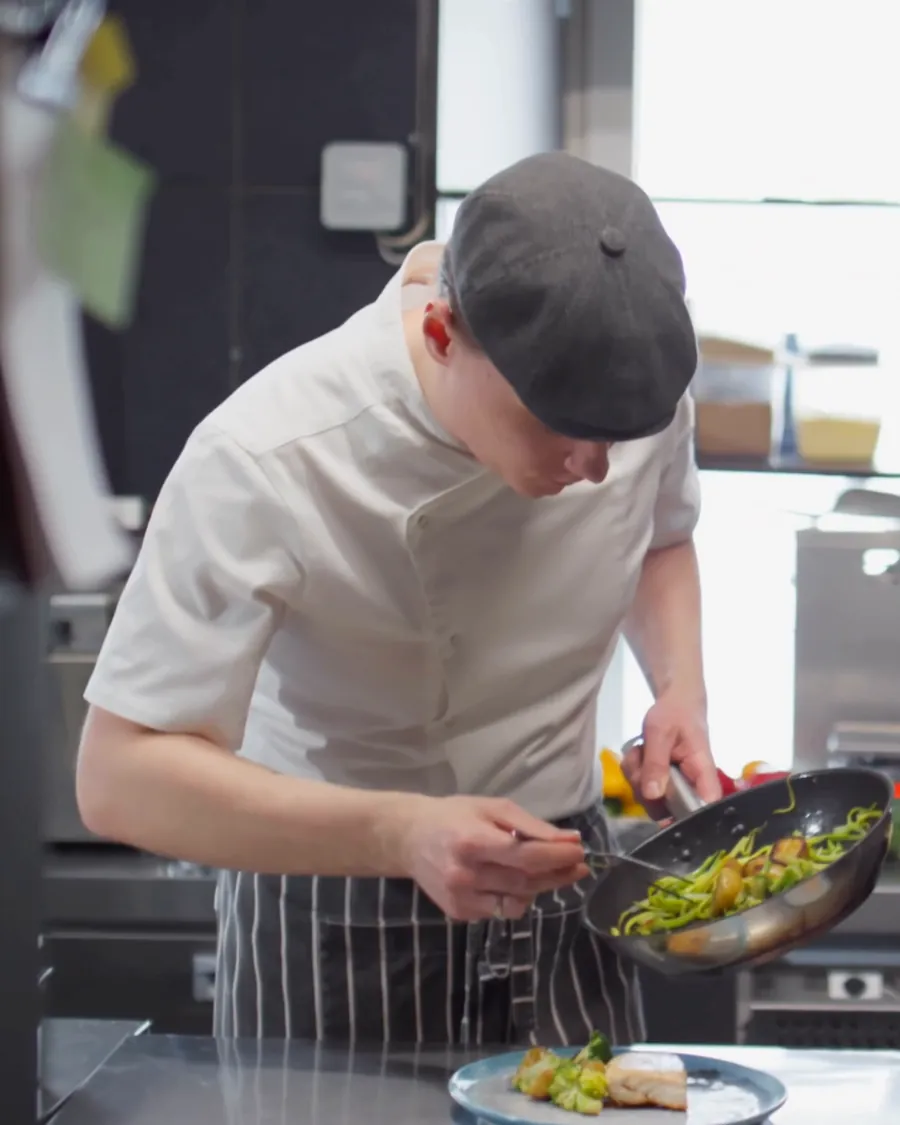 Chef in a cap tossing greens in a pan at a busy restaurant kitchen pass