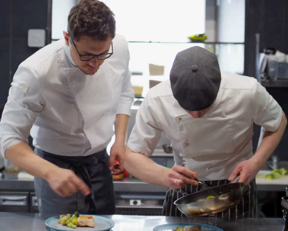 Two chefs in whites plating dishes side by side in a commercial kitchen